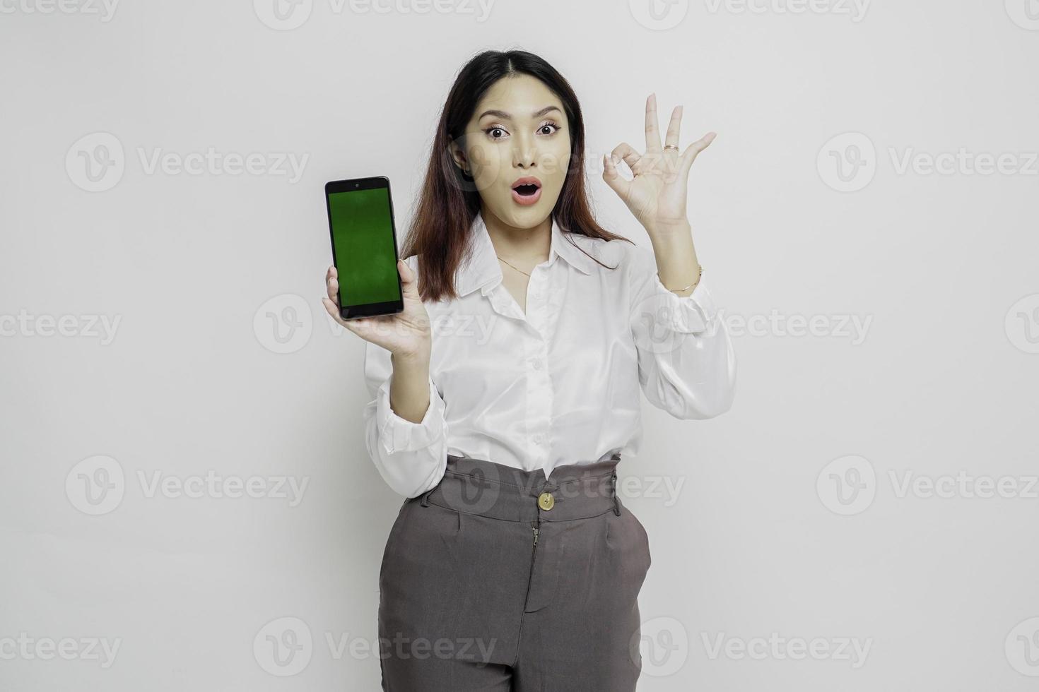 A portrait of a excited Asian woman wearing a white shirt while holding her phone and gesturing approval sign, isolated by white background photo