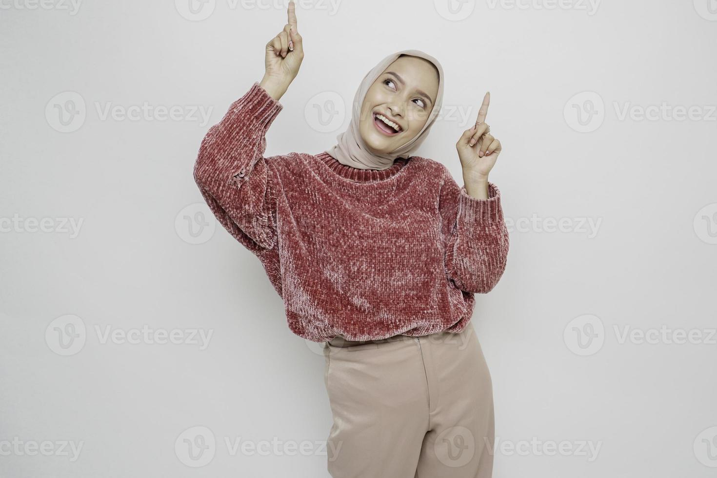 mujer asiática emocionada con suéter rosa y hiyab apuntando al espacio de la copia encima de ella, aislada de fondo blanco foto