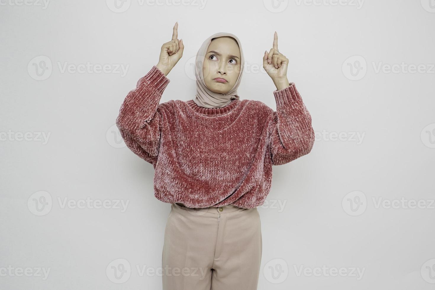 mujer musulmana asiática conmocionada con suéter rosa y hiyab señalando el espacio de copia encima de ella, aislada de fondo blanco foto