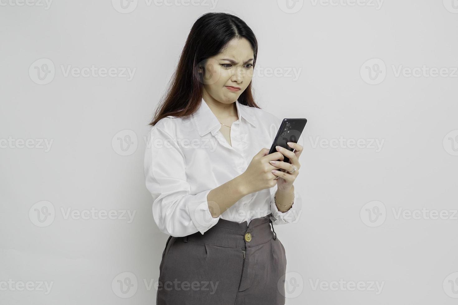 una joven asiática insatisfecha se ve descontenta usando camisa blanca expresiones faciales irritadas sosteniendo su teléfono foto