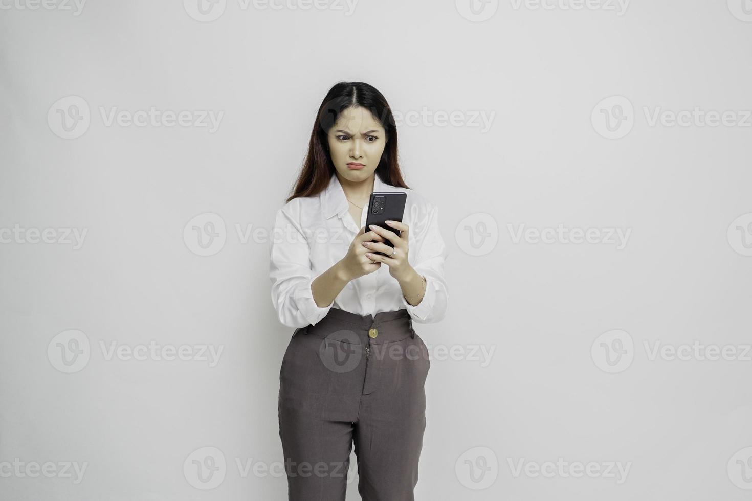 A dissatisfied young Asian woman looks disgruntled wearing white shirt irritated face expressions holding her phone photo