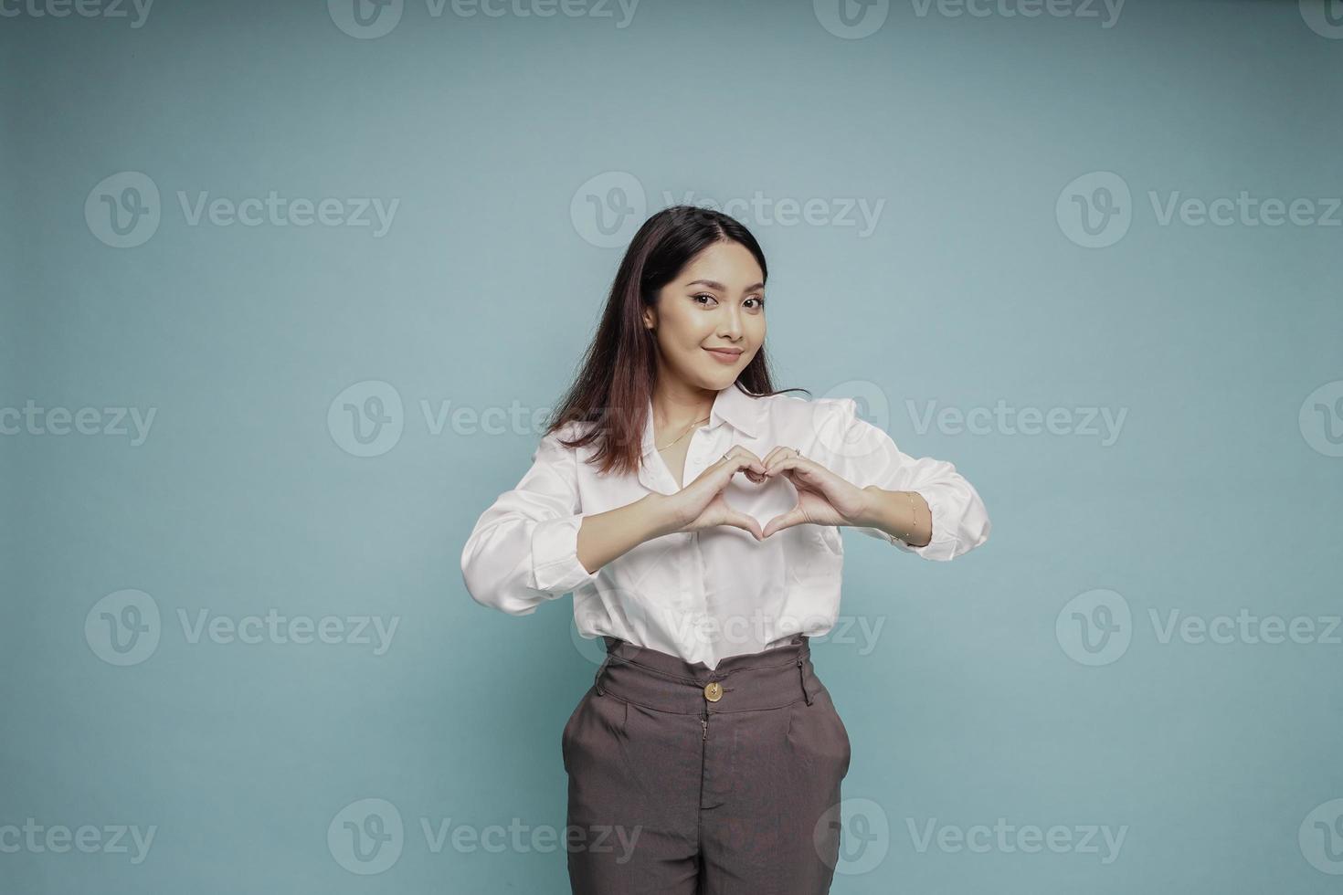 A happy young Asian woman wearing a white shirt feels romantic shapes heart gesture expresses tender feelings photo