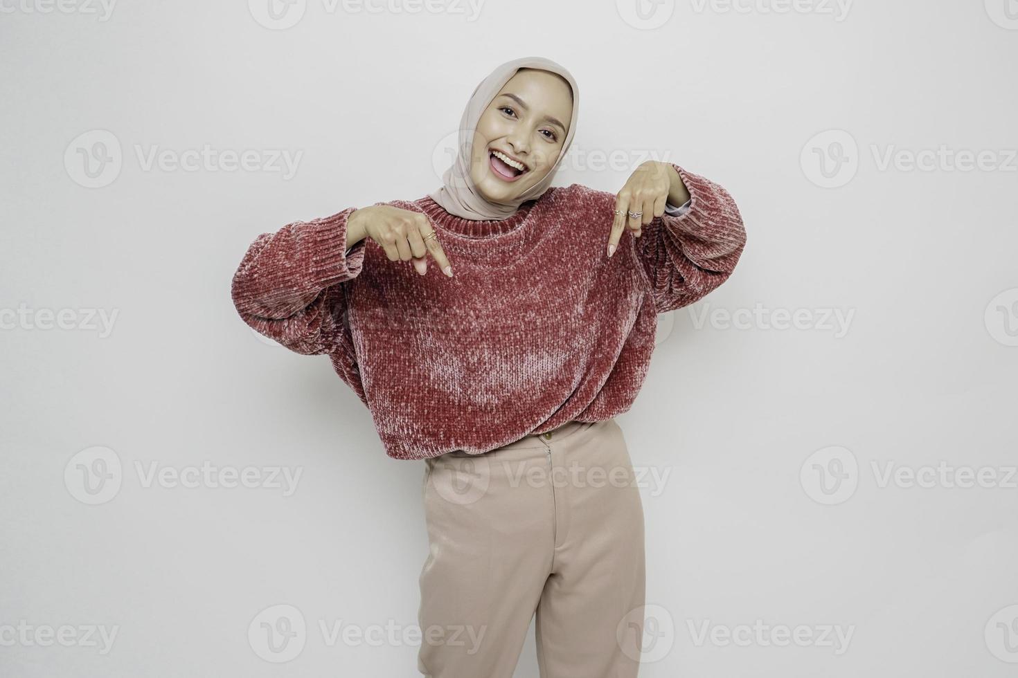 Excited Asian woman wearing pink sweater and hijab pointing at the copy space below her, isolated by white background photo