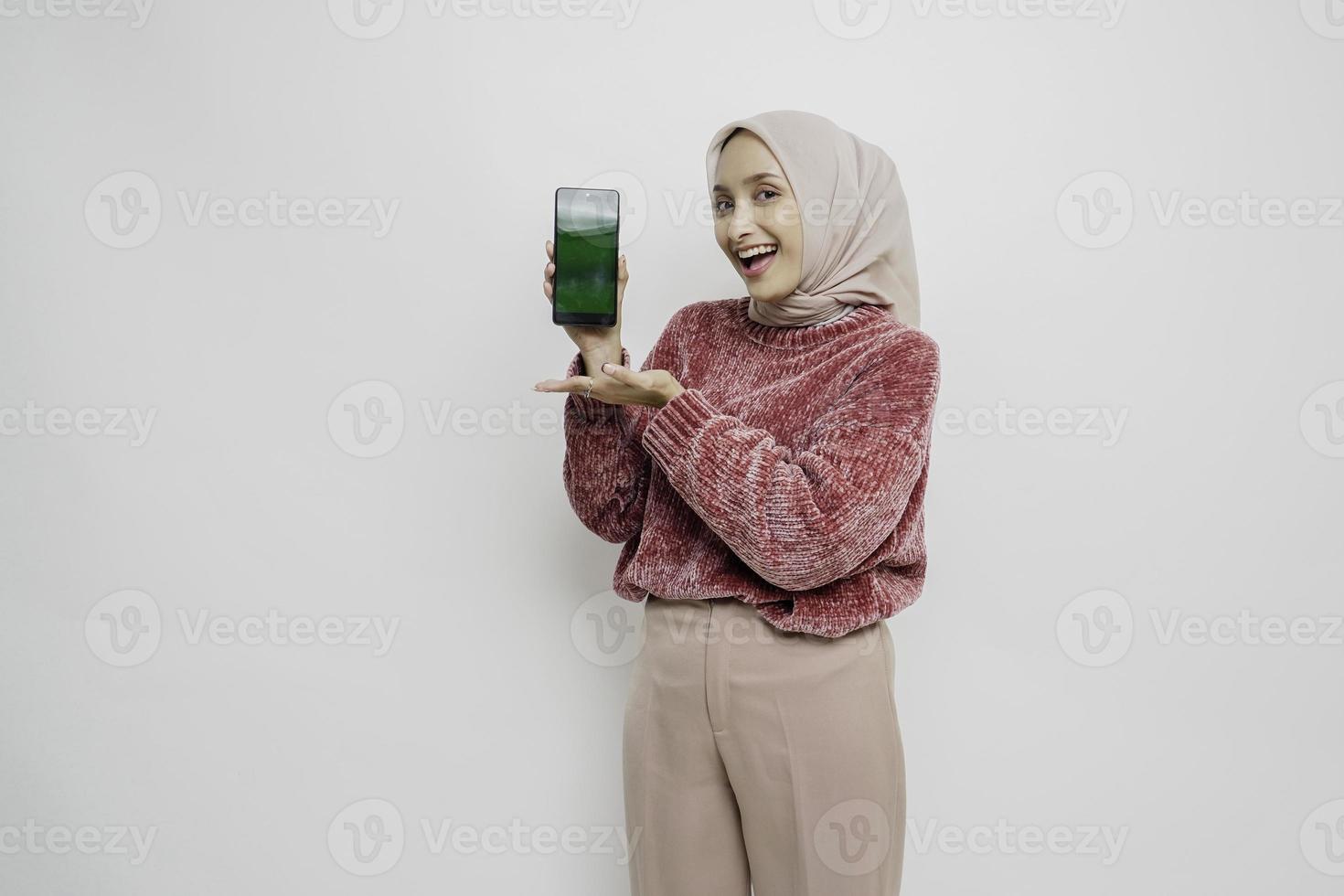 Excited Asian Muslim woman wearing pink sweater and hijab pointing at the copy space beside her while holding her phone, isolated by white background photo