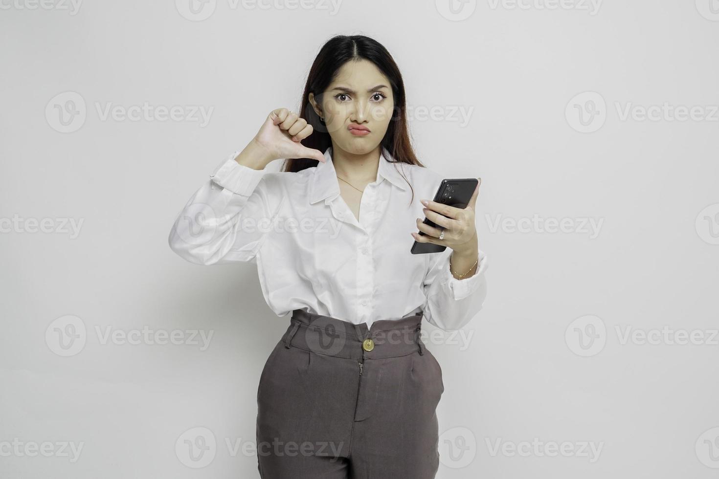 una mujer asiática decepcionada con pantalones blanco da un gesto de desaprobación con la mano hacia abajo, aislada de fondo blanco foto