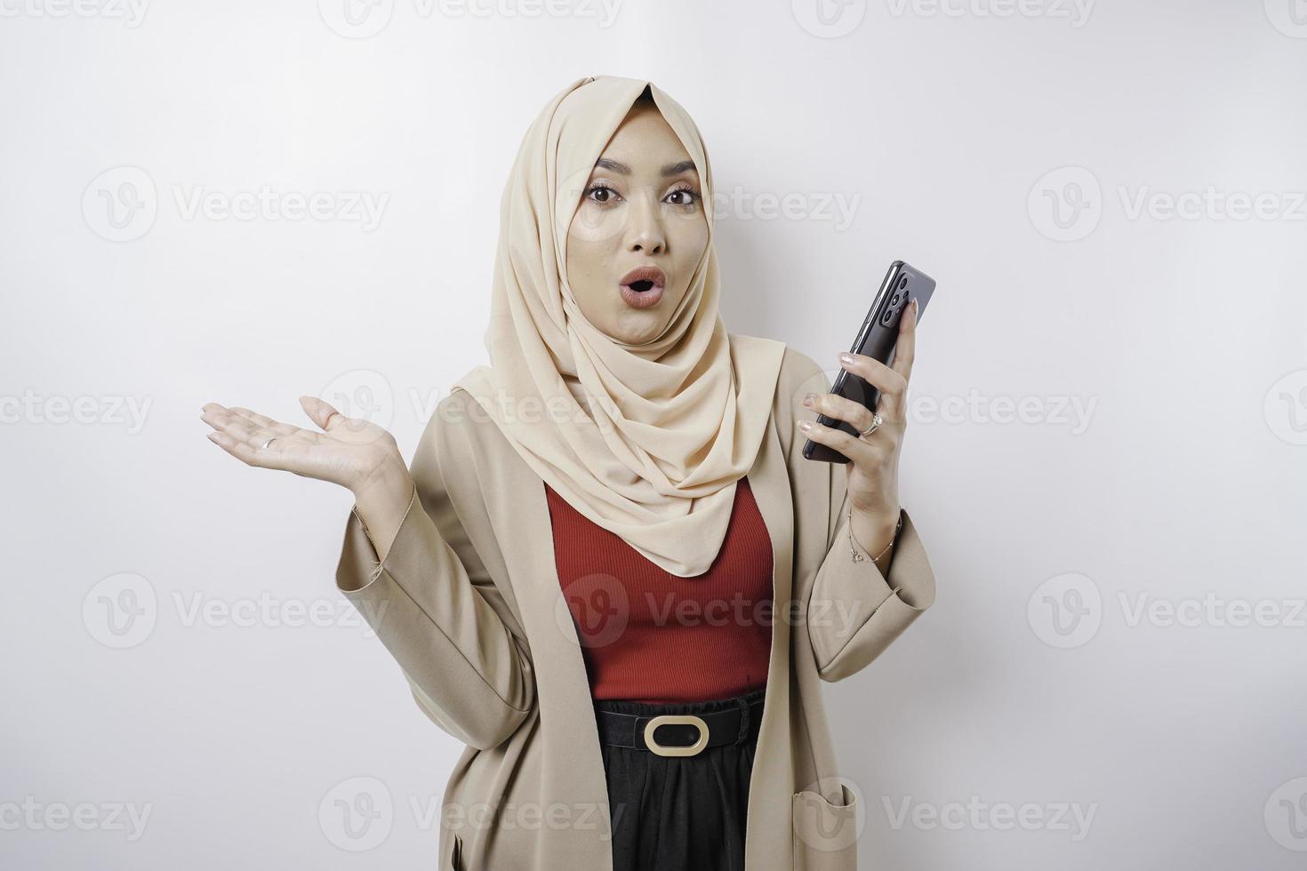 Shocked Asian woman wearing hijab pointing at the copy space beside her while holding her phone, isolated by white background photo