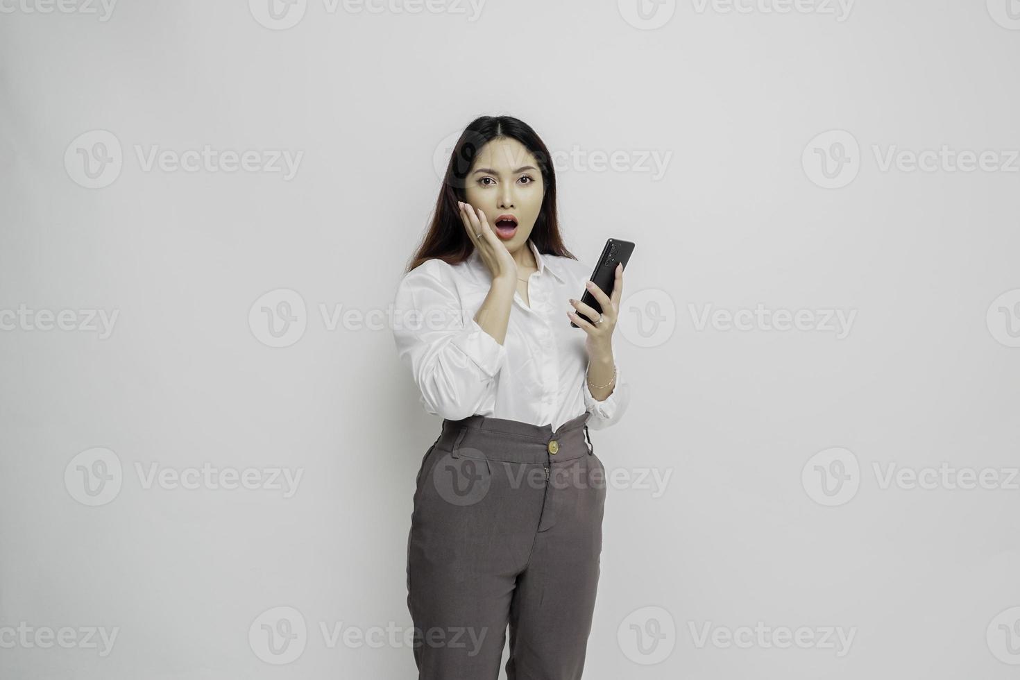 Shocked Asian woman wearing white shirt and holding her phone, isolated by white background photo
