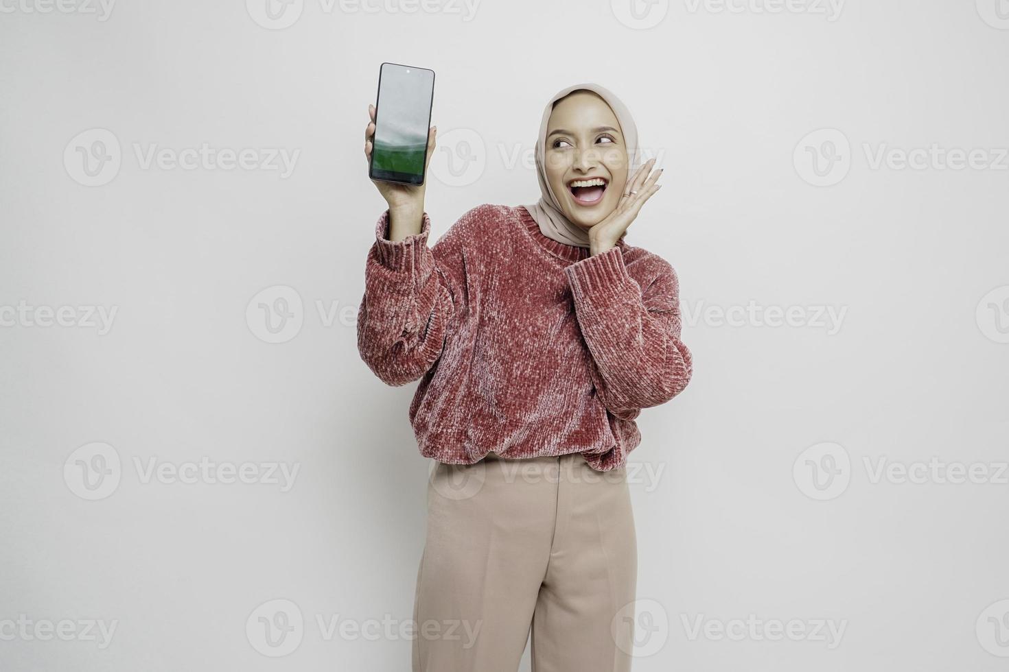 mujer musulmana asiática emocionada con suéter rosa y hiyab señalando el espacio de copia a su lado mientras sostiene su teléfono, aislada por fondo blanco foto