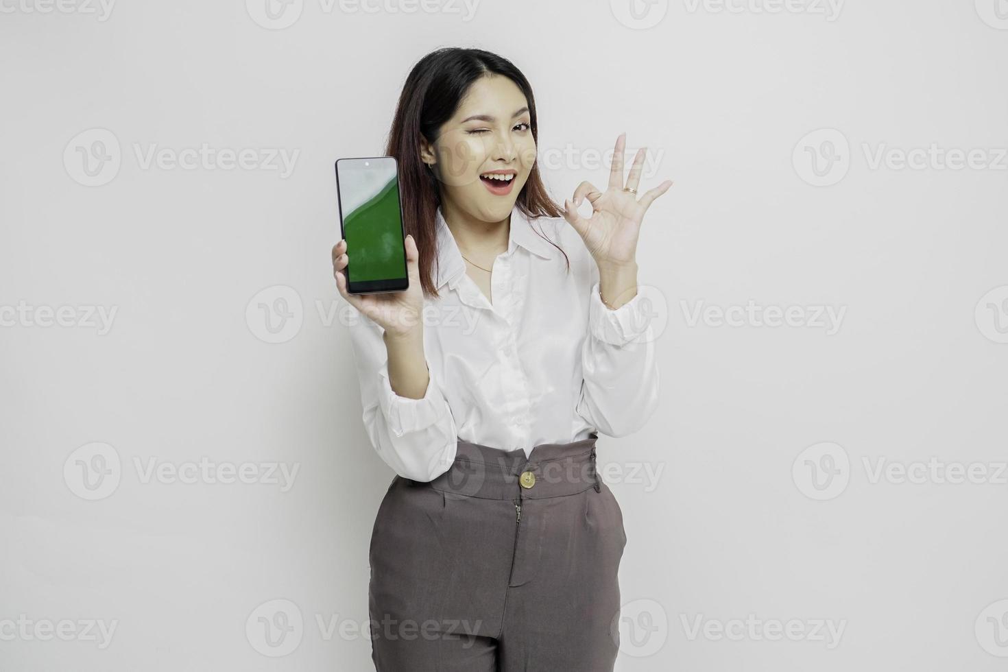 A portrait of a excited Asian woman wearing a white shirt while holding her phone and gesturing approval sign, isolated by white background photo