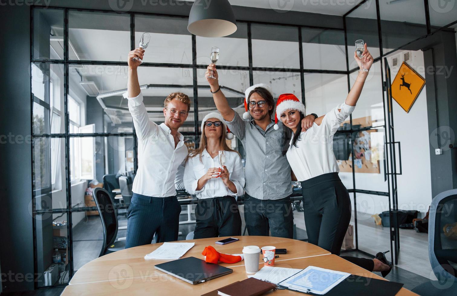 With hands up. Employees in formal clothes, glasses with champagne and in christmas hats celebrating new year in the office photo