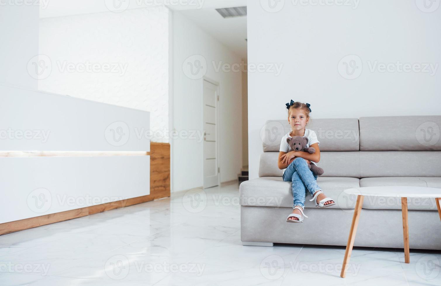 Cute little girl with teddy bear in hands sits in waiting room of hospital photo