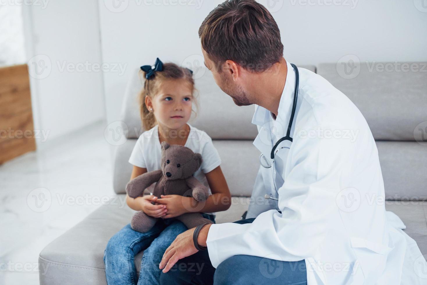 Male doctor in white uniform sits in the clinic with little girl photo