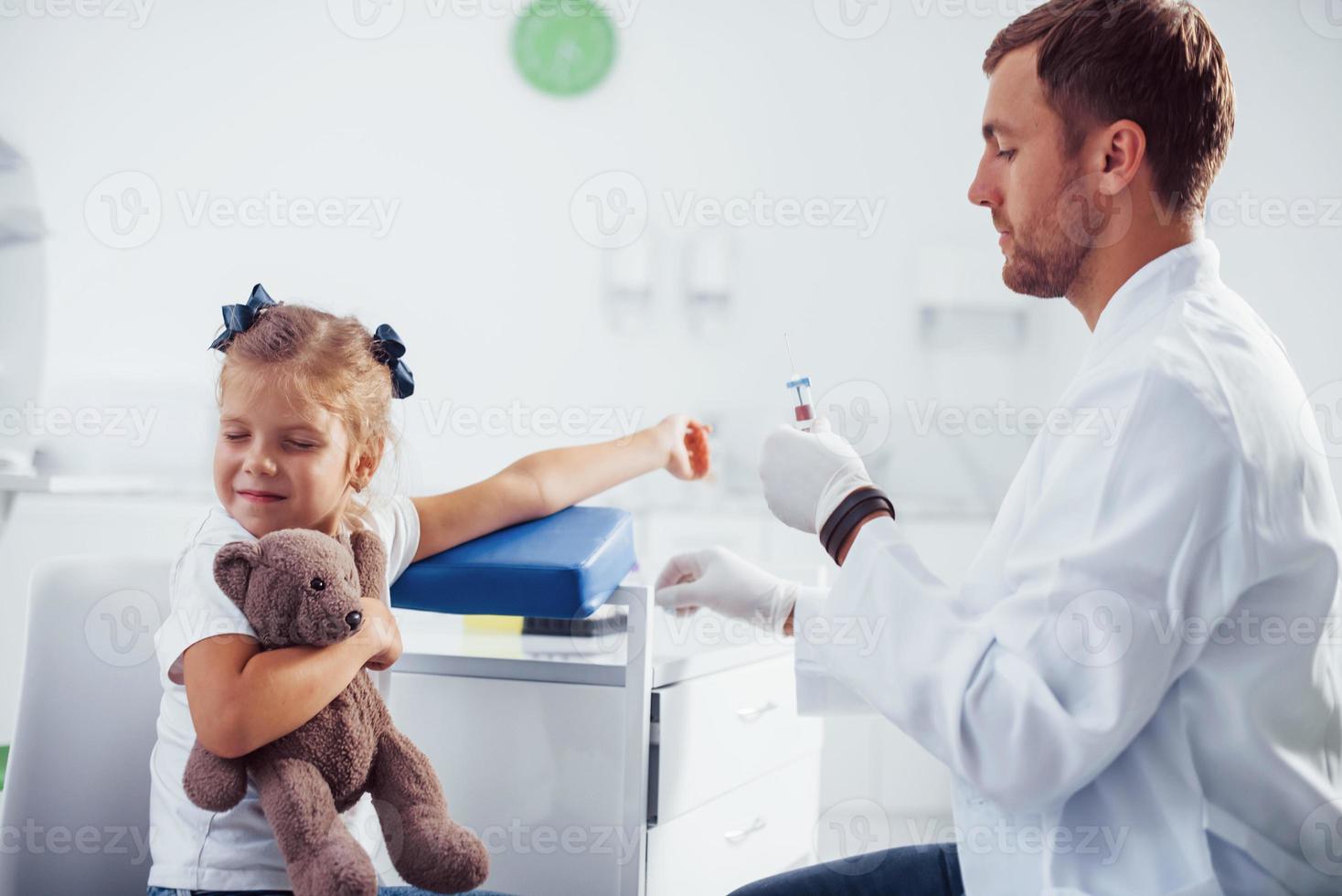 muestra de sangre. niña con su osito de peluche está en la clínica con el médico foto