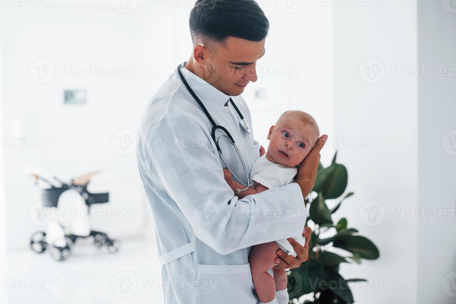 Stands against window. Young pediatrician is with little baby in the clinic at daytime photo