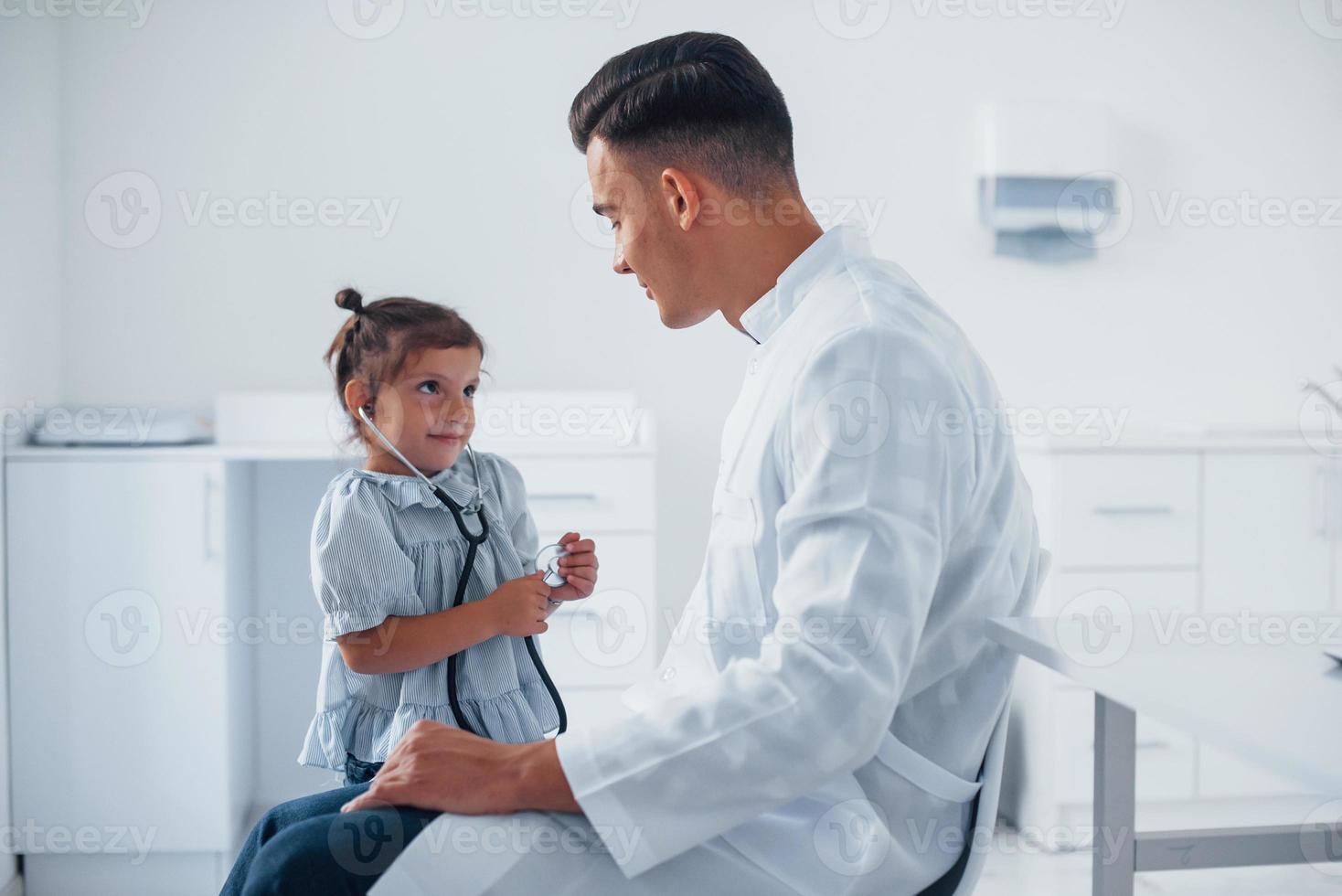 Teaches girl how to use stethoscope. Young pediatrician works with little female visitor in the clinic photo