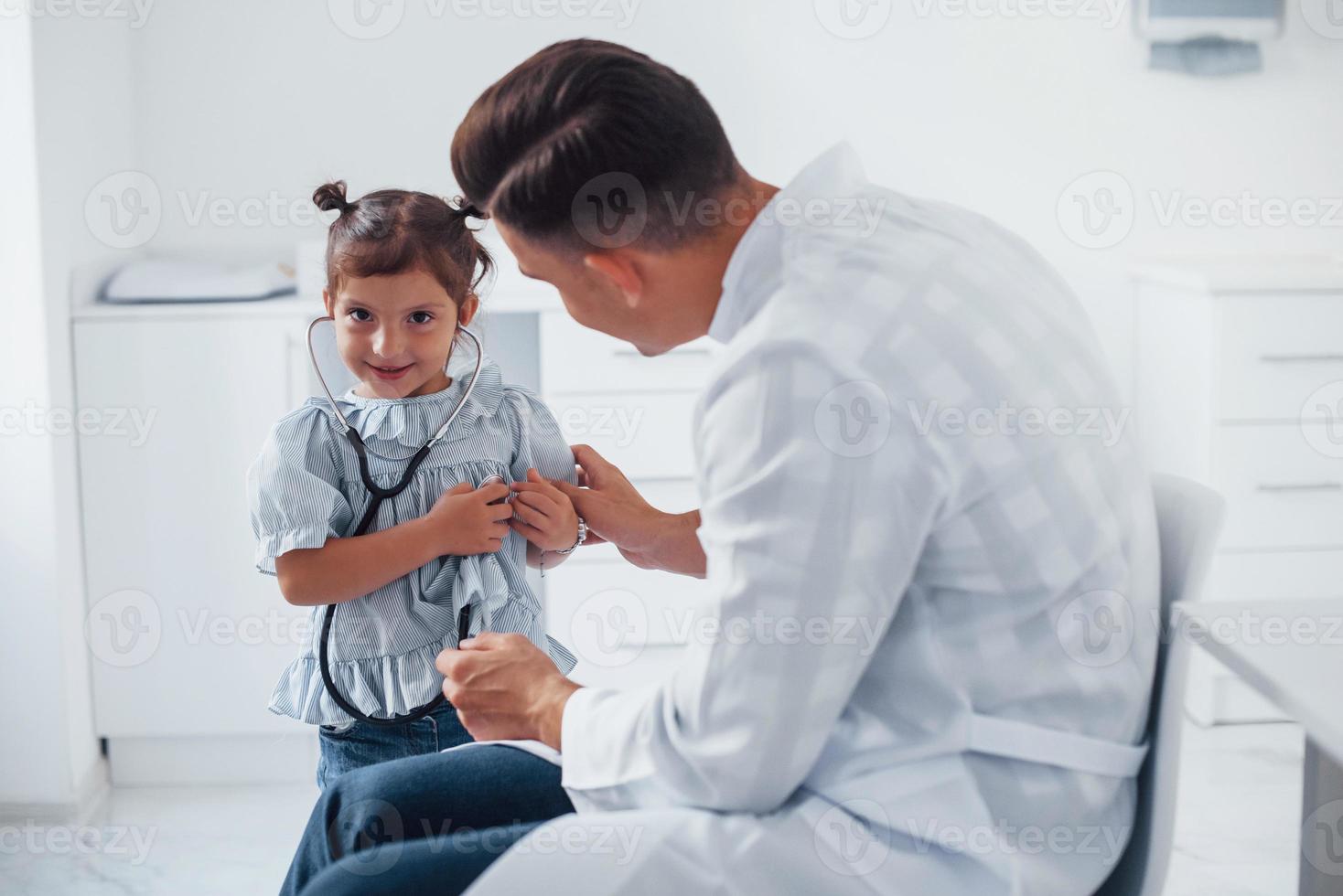 enseña a la niña a usar el estetoscopio. joven pediatra trabaja con una pequeña visitante en la clínica foto