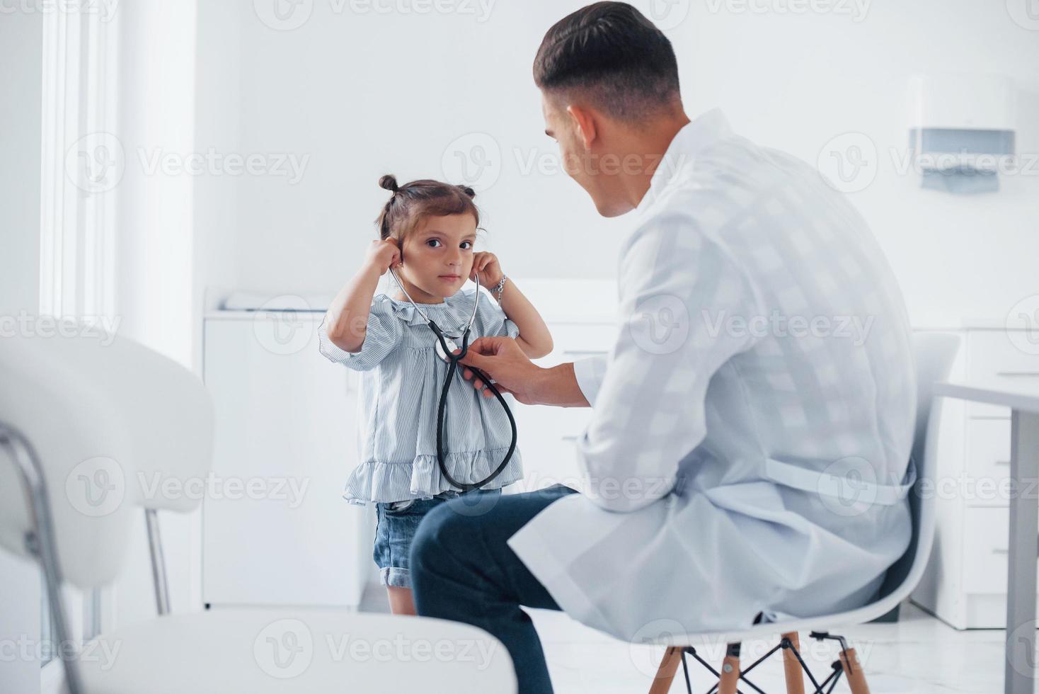 enseña a la niña a usar el estetoscopio. joven pediatra trabaja con una pequeña visitante en la clínica foto