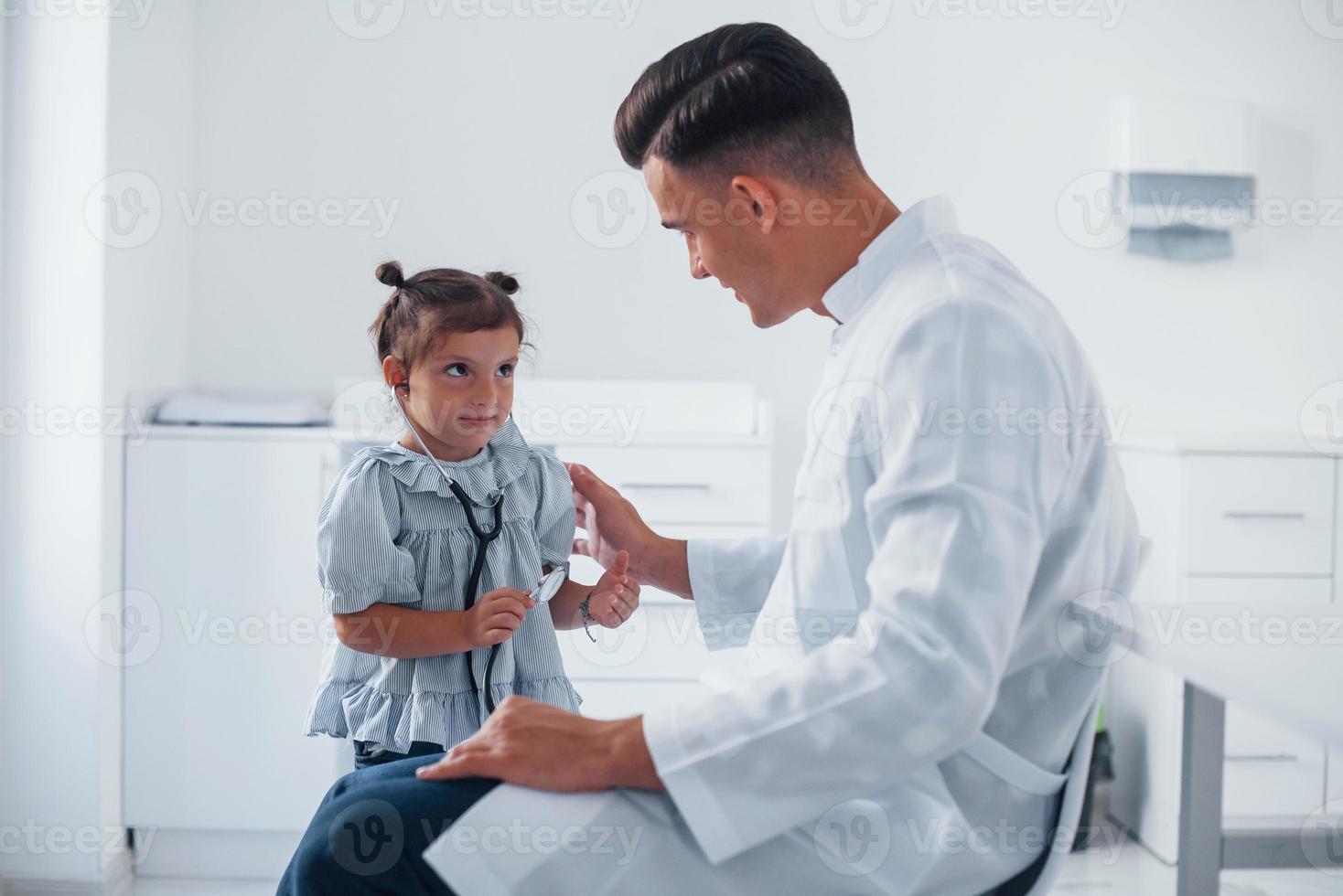 Teaches girl how to use stethoscope. Young pediatrician works with little female visitor in the clinic photo