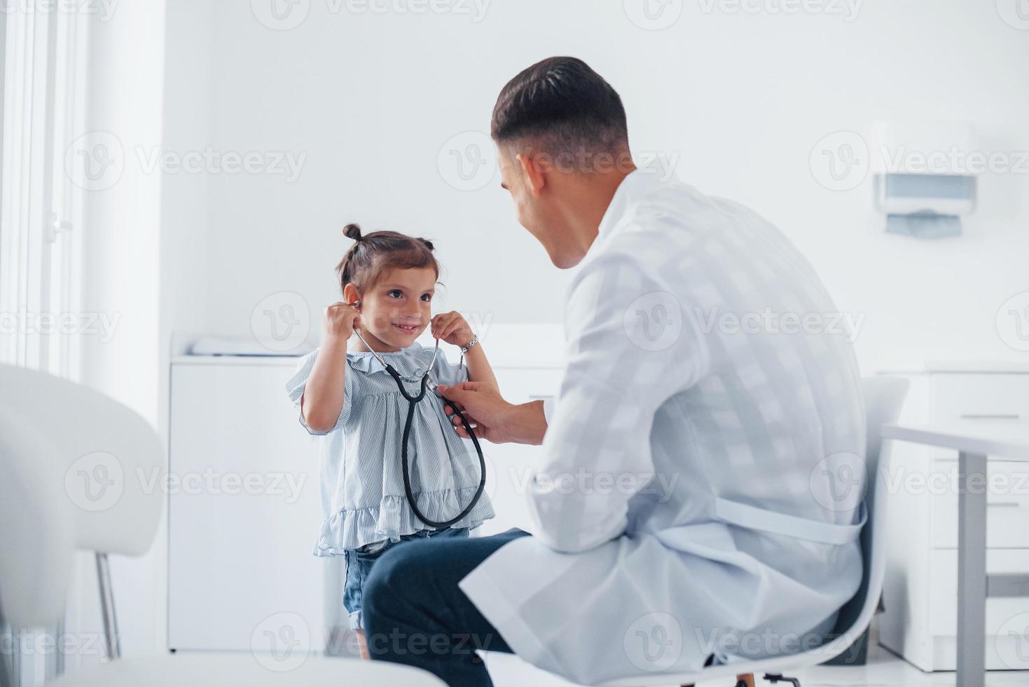 Teaches girl how to use stethoscope. Young pediatrician works with little female visitor in the clinic photo