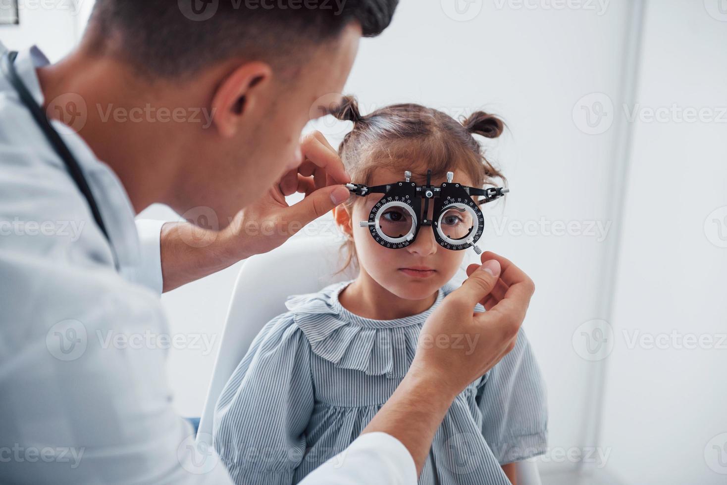 probando la visión. joven oftalmólogo está con una pequeña visitante en la clínica foto