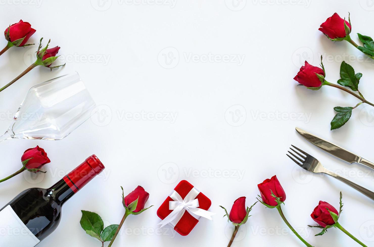 comedor de san valentín sobre fondo blanco con caja de regalo roja, una botella de vino tinto, vaso, cuchillo, tenedor y rosas rojas. foto