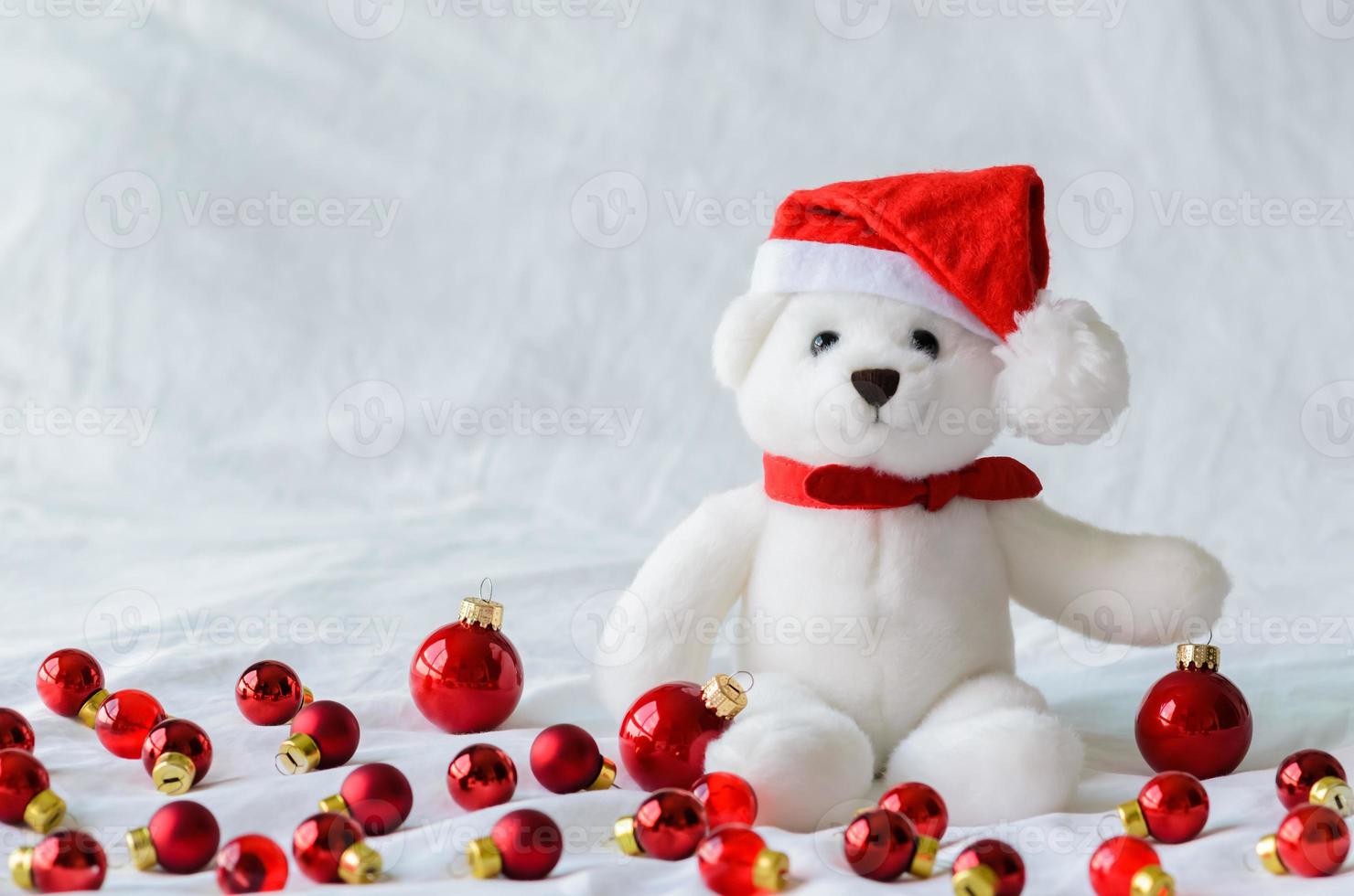 enfoque selectivo en los ojos de oso de peluche de santa claus que usan sombrero sentado con adornos navideños rojos sobre fondo de tela blanca. foto