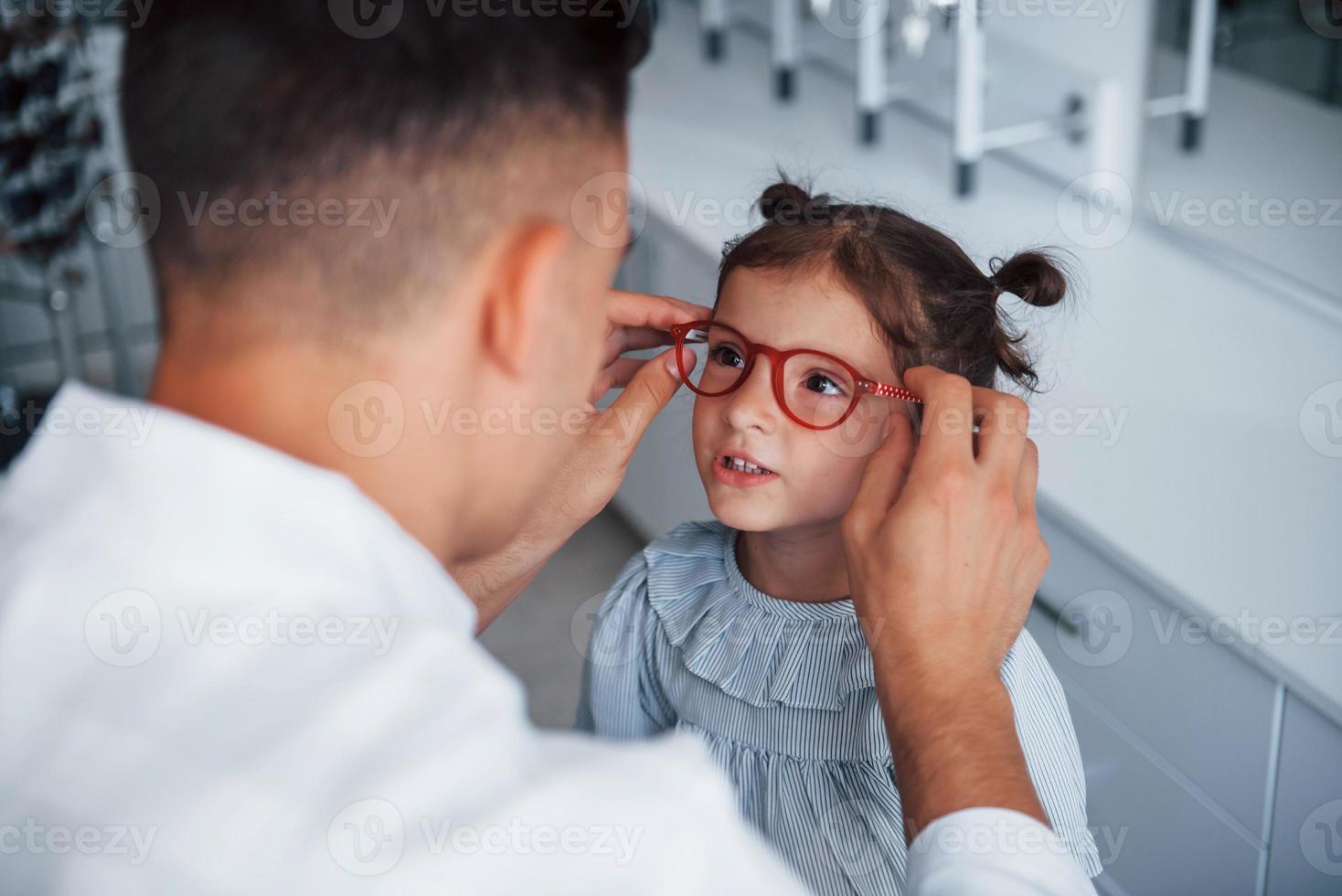 Young pediatrician in white coat helps to get new glasses for little girl photo