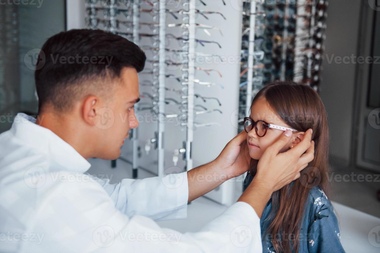 Young pediatrician in white coat helps to get new glasses for little girl photo