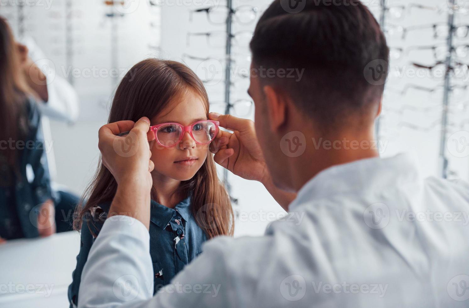 Young pediatrician in white coat helps to get new glasses for little girl photo