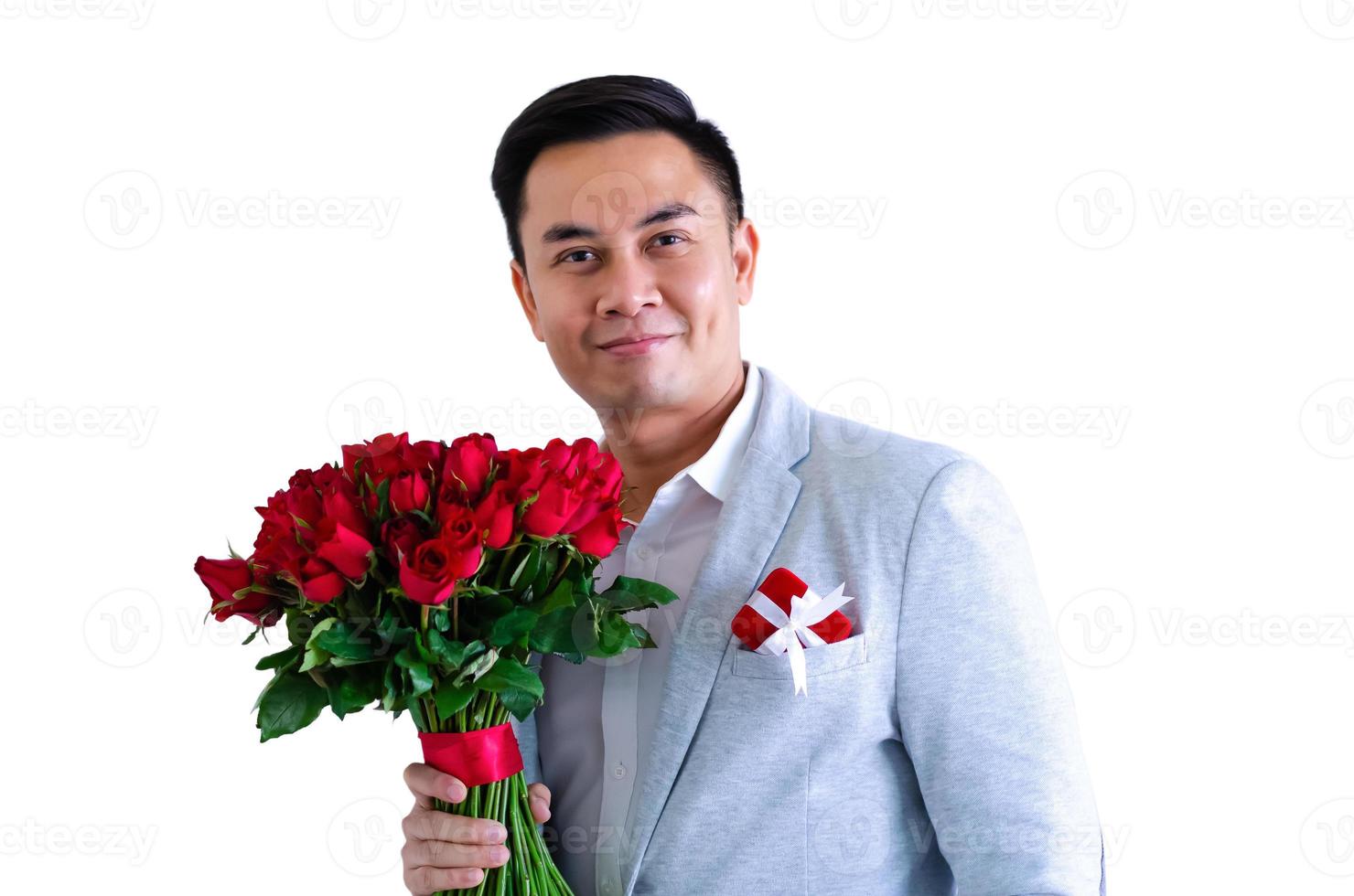 Asian man wearing grey suit holding a bouquet of red roses and red gift box isolated in white background for anniversary or Valentine day concept. photo