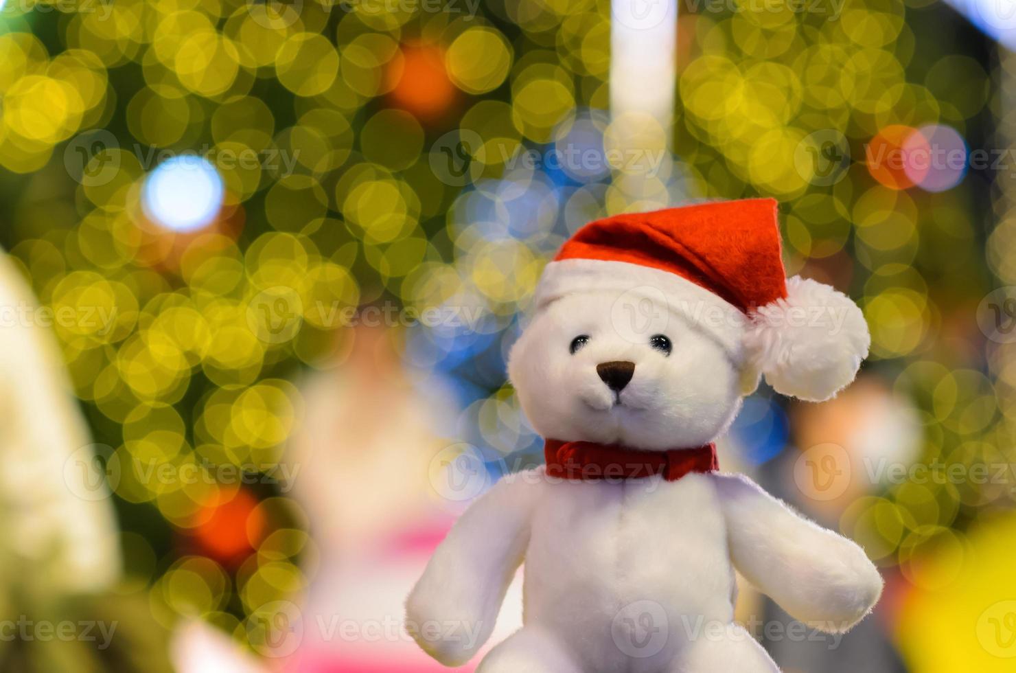 Santa claus teddy bear wearing hat sitting in front of colorful bokeh lights of Christmas tree. photo