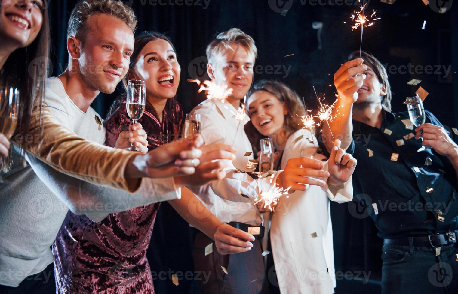 Having fun with sparklers. Confetti is in the air. Group of cheerful friends celebrating new year indoors with drinks in hands photo