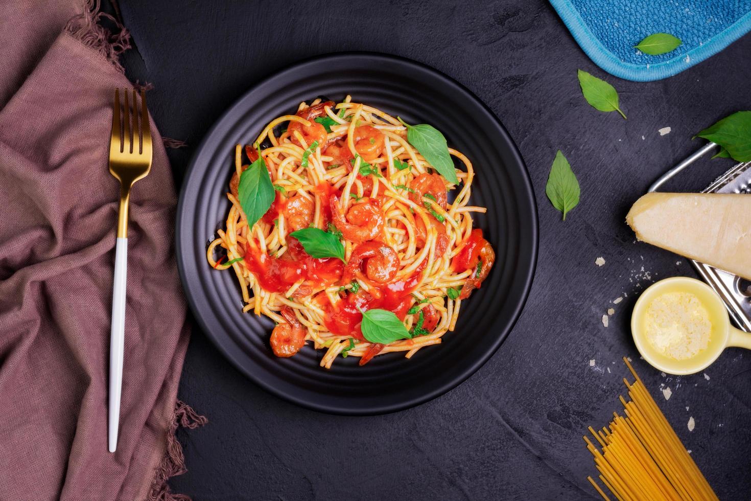 Delicious spaghetti pasta with prawns and cheese served on a black plate. With vegetables, Italian tomato sauce, and spices arranged on a wooden table, black background, top view photo