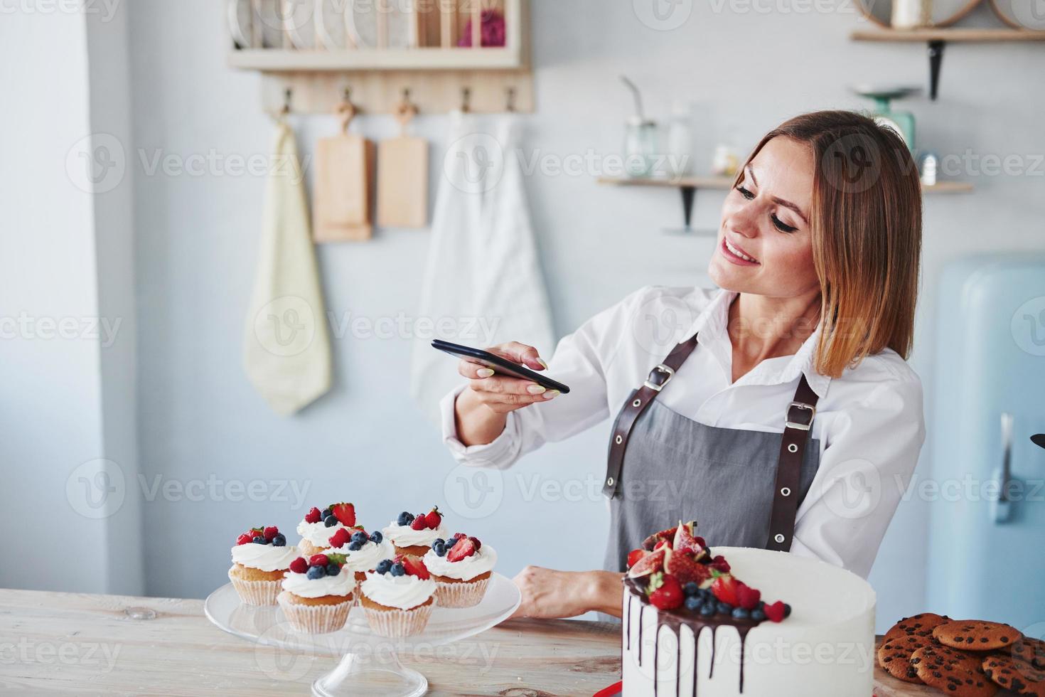 Woman stands in the kitchen and takes photo of her homemade cookies and pie by using phone