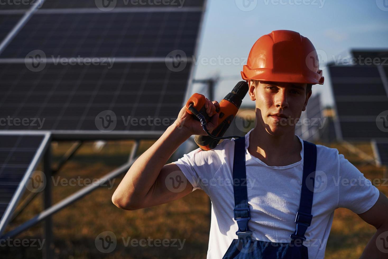 Using cordless screwdriver. Male worker in blue uniform outdoors with solar batteries at sunny day photo