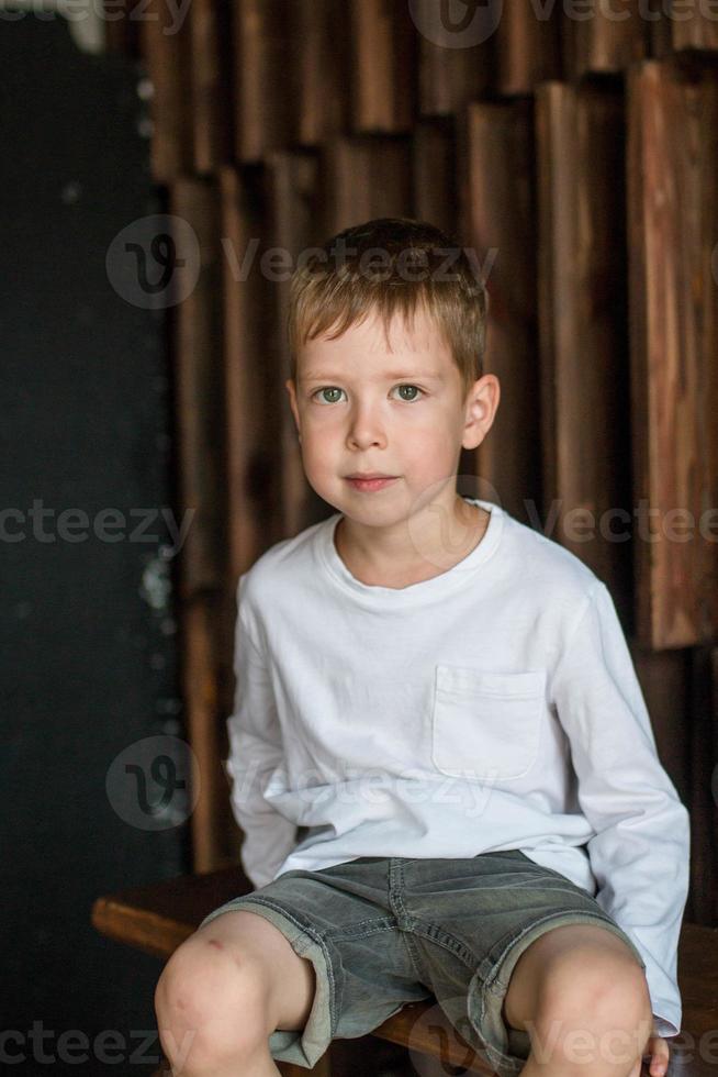 portrait of a little blond boy in a white t-shirt sits against the background of a wooden wall and smiles, looks at the camera. photo