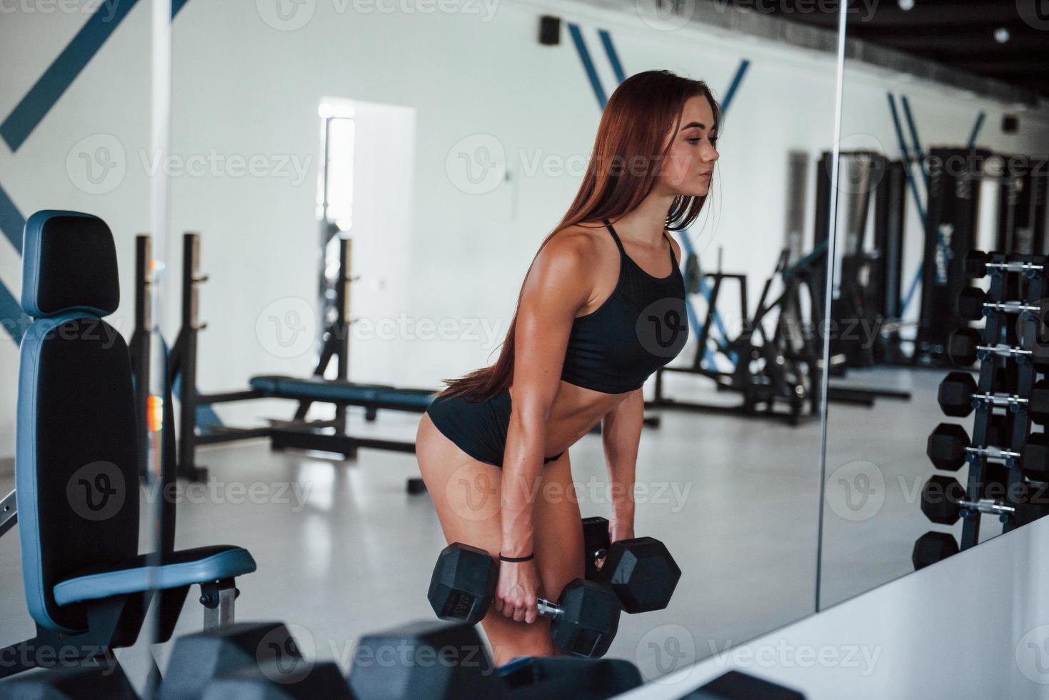 Young fitness woman is in the gym near mirror with dumbbells in hands photo