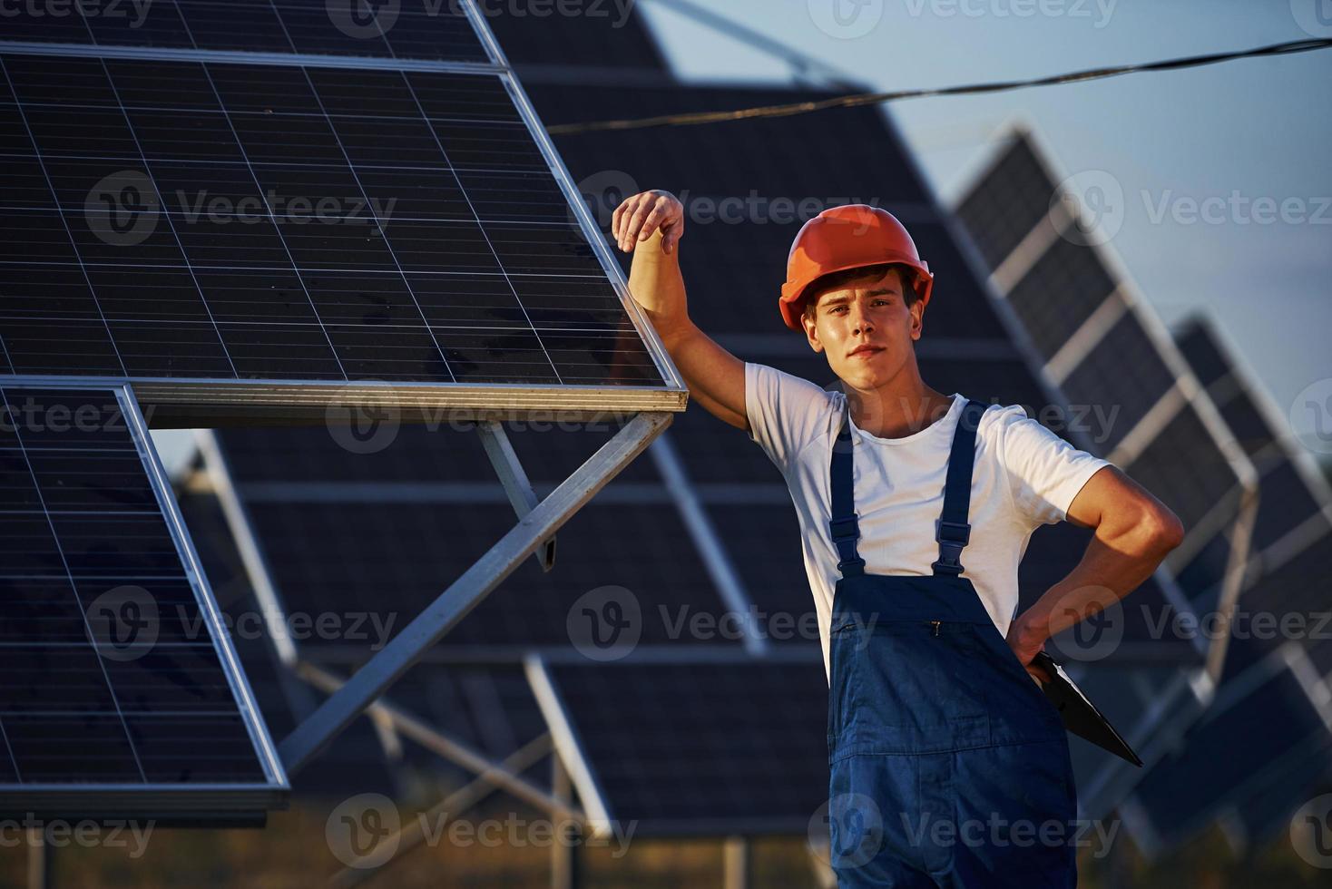 Holds notepad in hand. Male worker in blue uniform outdoors with solar batteries at sunny day photo