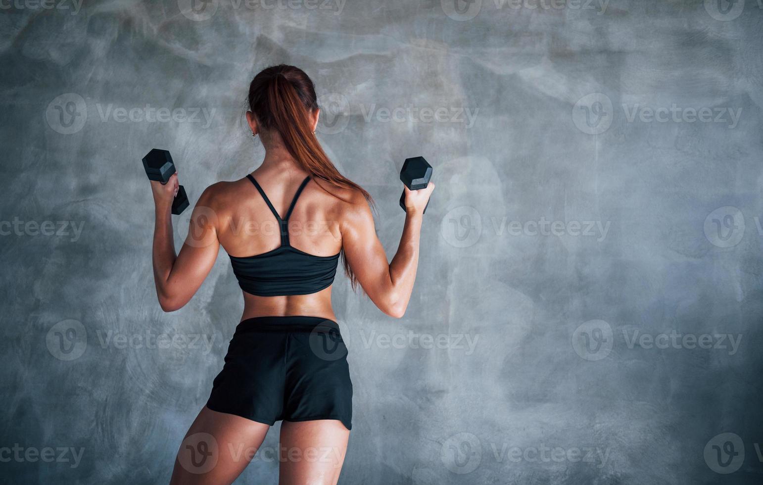 Young fitness woman is in the gym near wall with dumbbells in hands photo