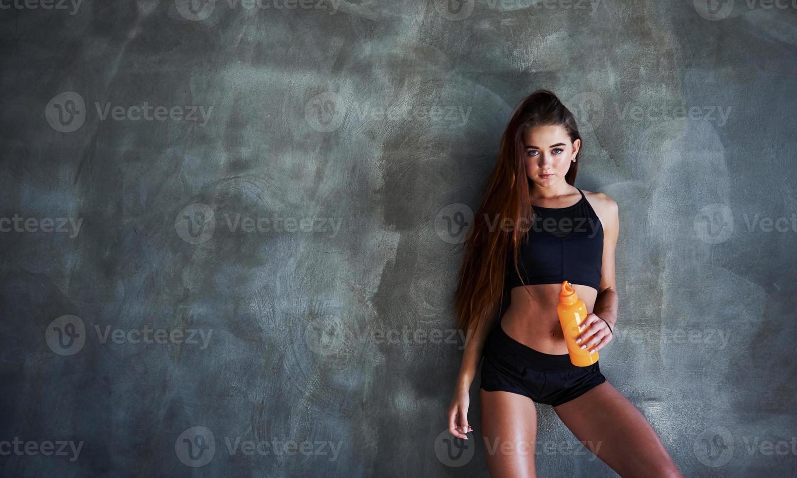 Young fitness woman with slim type of body and bottle of water is leaning on the wall photo