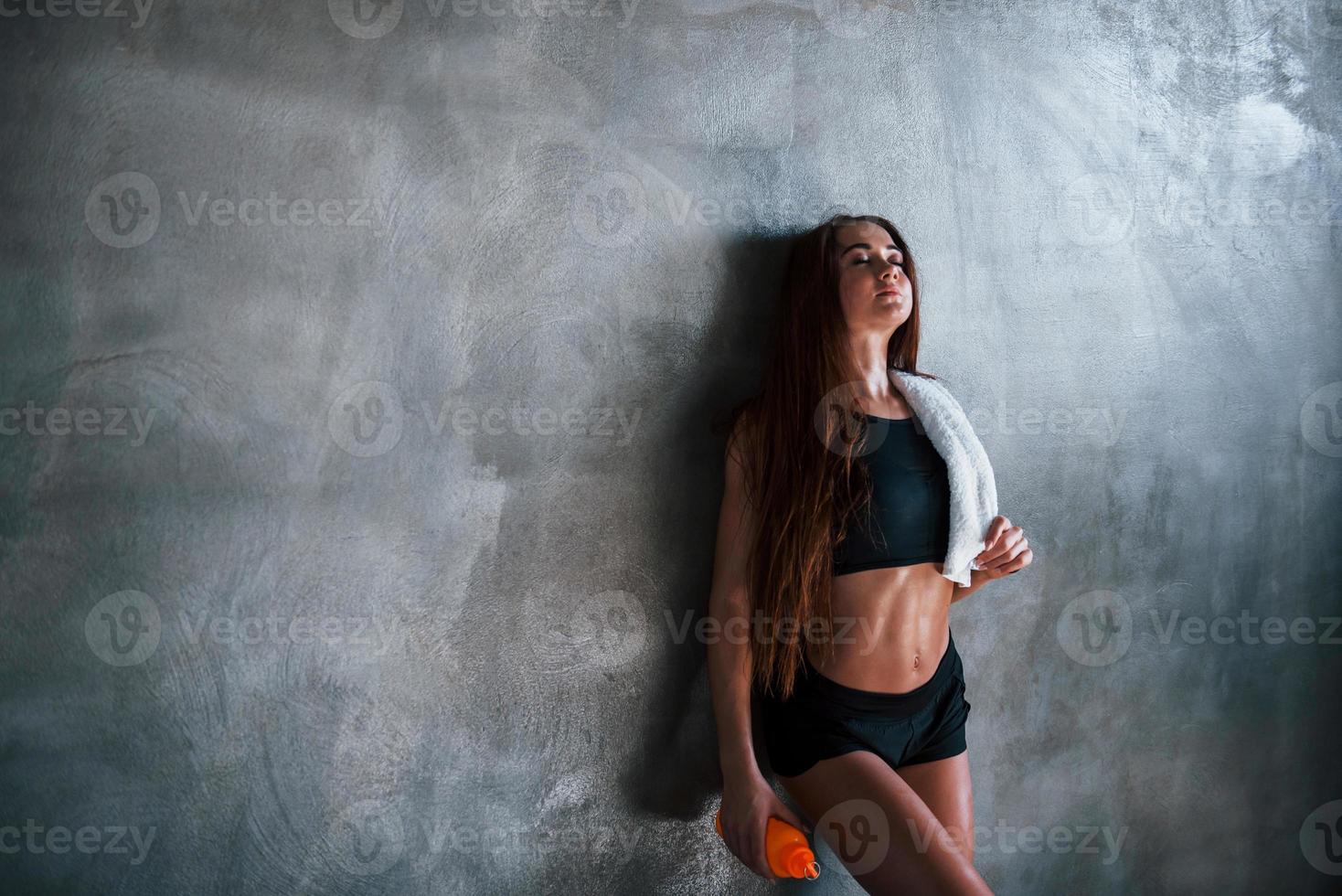 Young fitness woman with slim type of body and bottle of water is leaning on the wall photo
