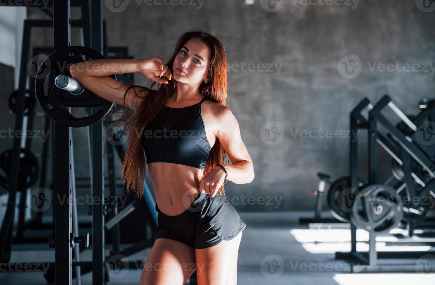 posando para la cámara. una joven deportista con un tipo de cuerpo delgado y ropa deportiva negra está en el gimnasio foto