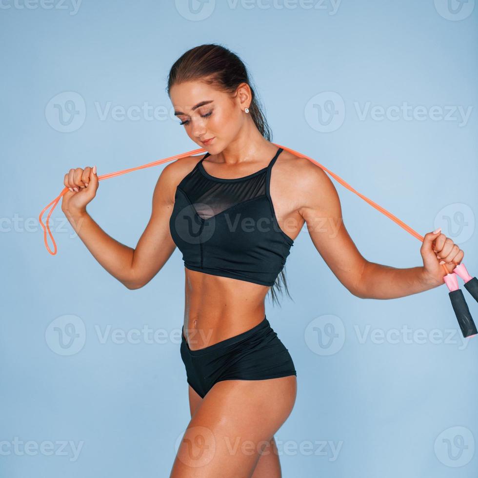 Stands with jumping rope. Young woman with slim body type isolated against blue background photo