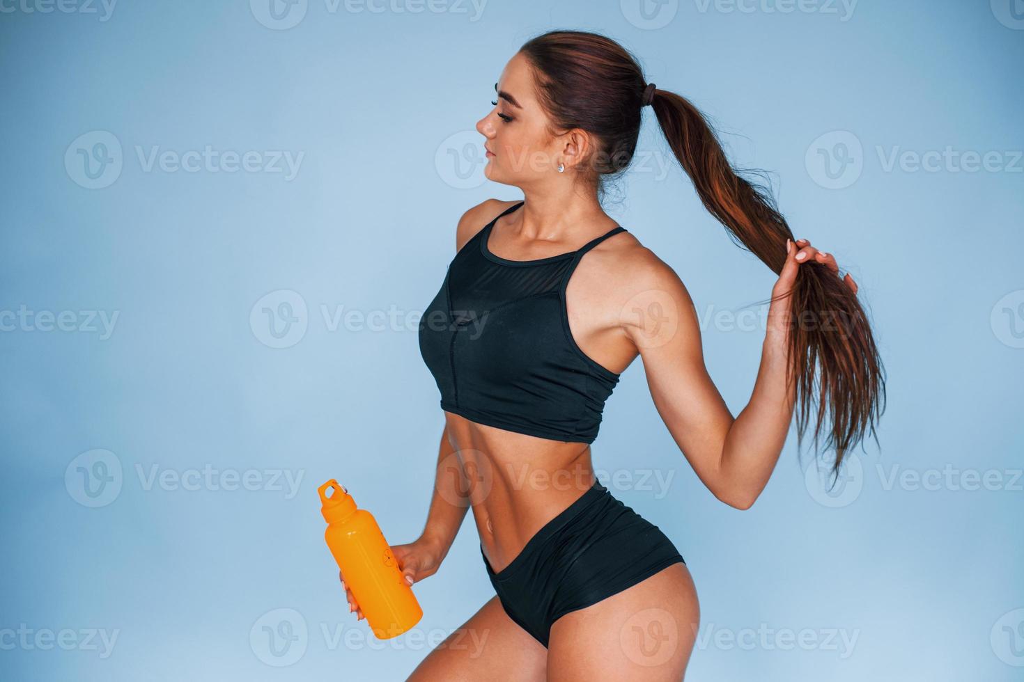 Takes a break and holds bottle of water. Young woman with slim body type isolated against blue background photo