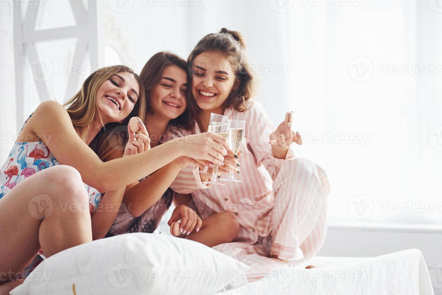 Celebrating with glasses of alcohol in hands. Happy female friends having good time at pajama party in the bedroom photo