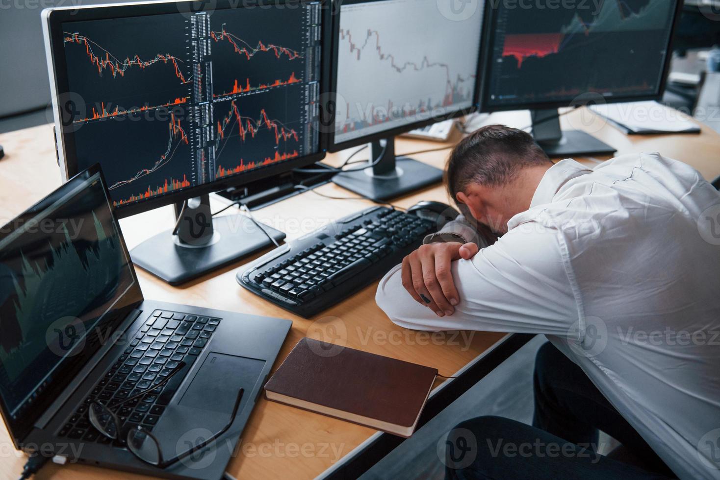 Tired businessman sleeping on the workplace by leaning on the table with multiple screens on it. Stock information on displays photo