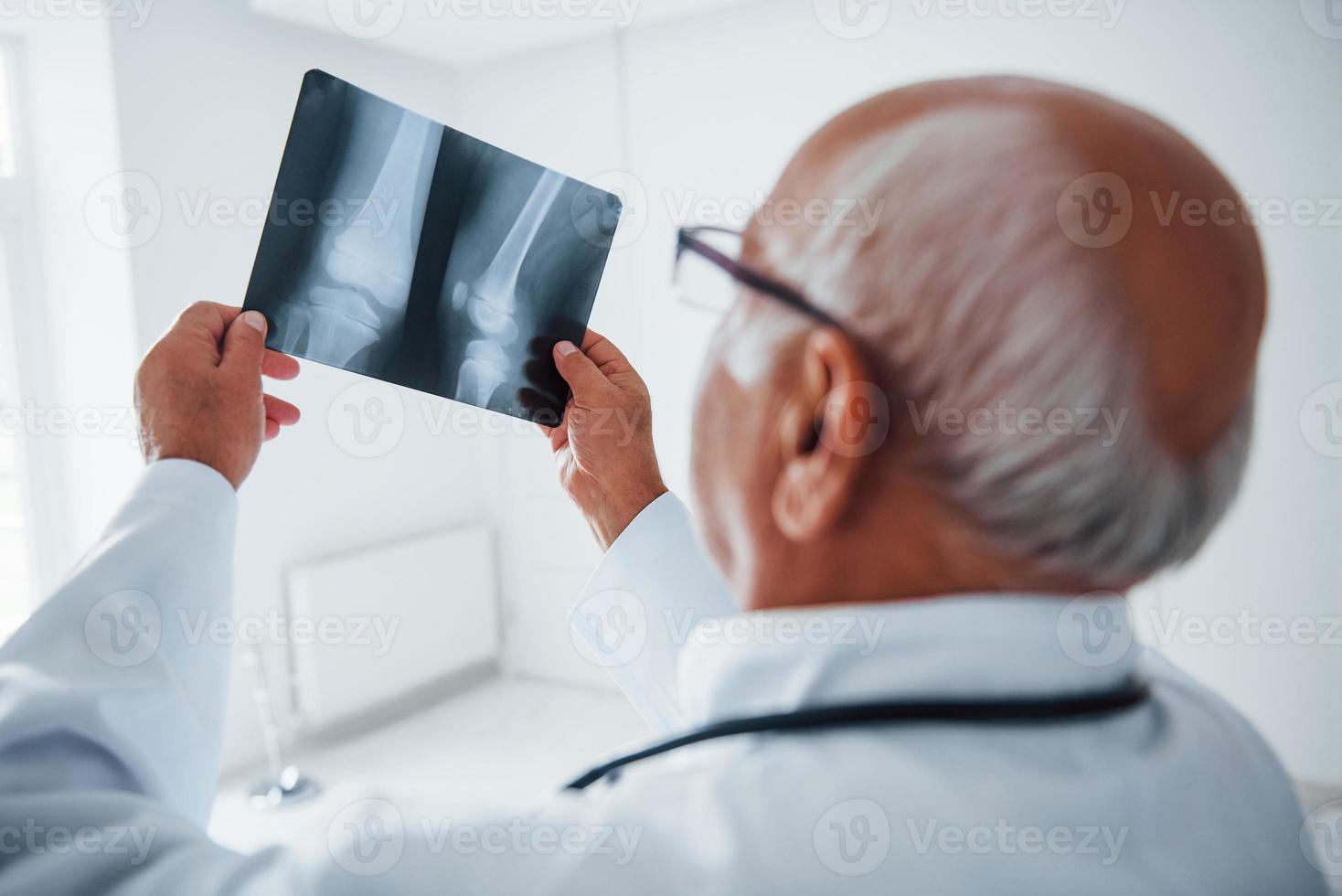 médico senior en uniforme blanco examina la radiografía de las piernas humanas foto