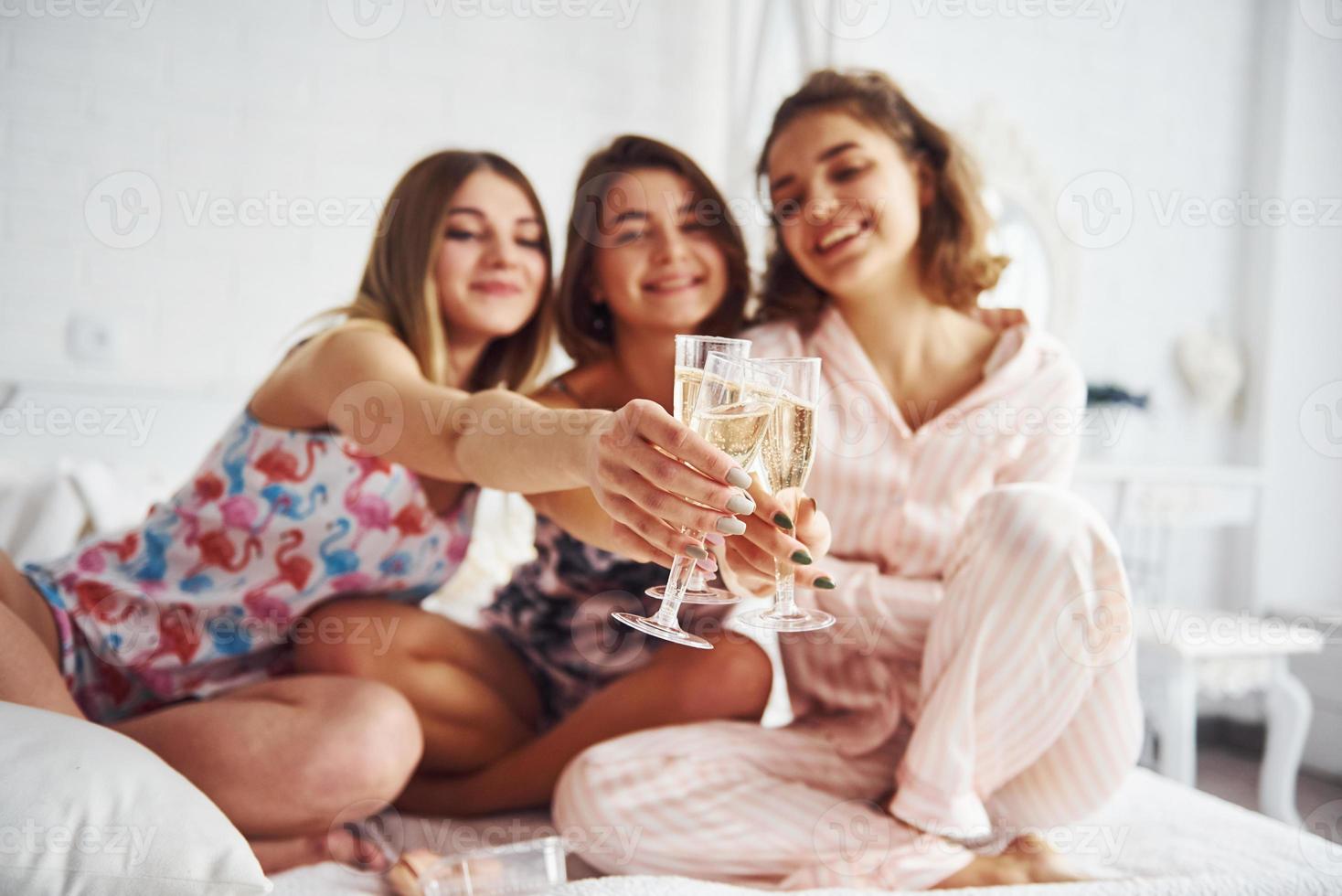 Celebrating with glasses of alcohol in hands. Happy female friends having good time at pajama party in the bedroom photo