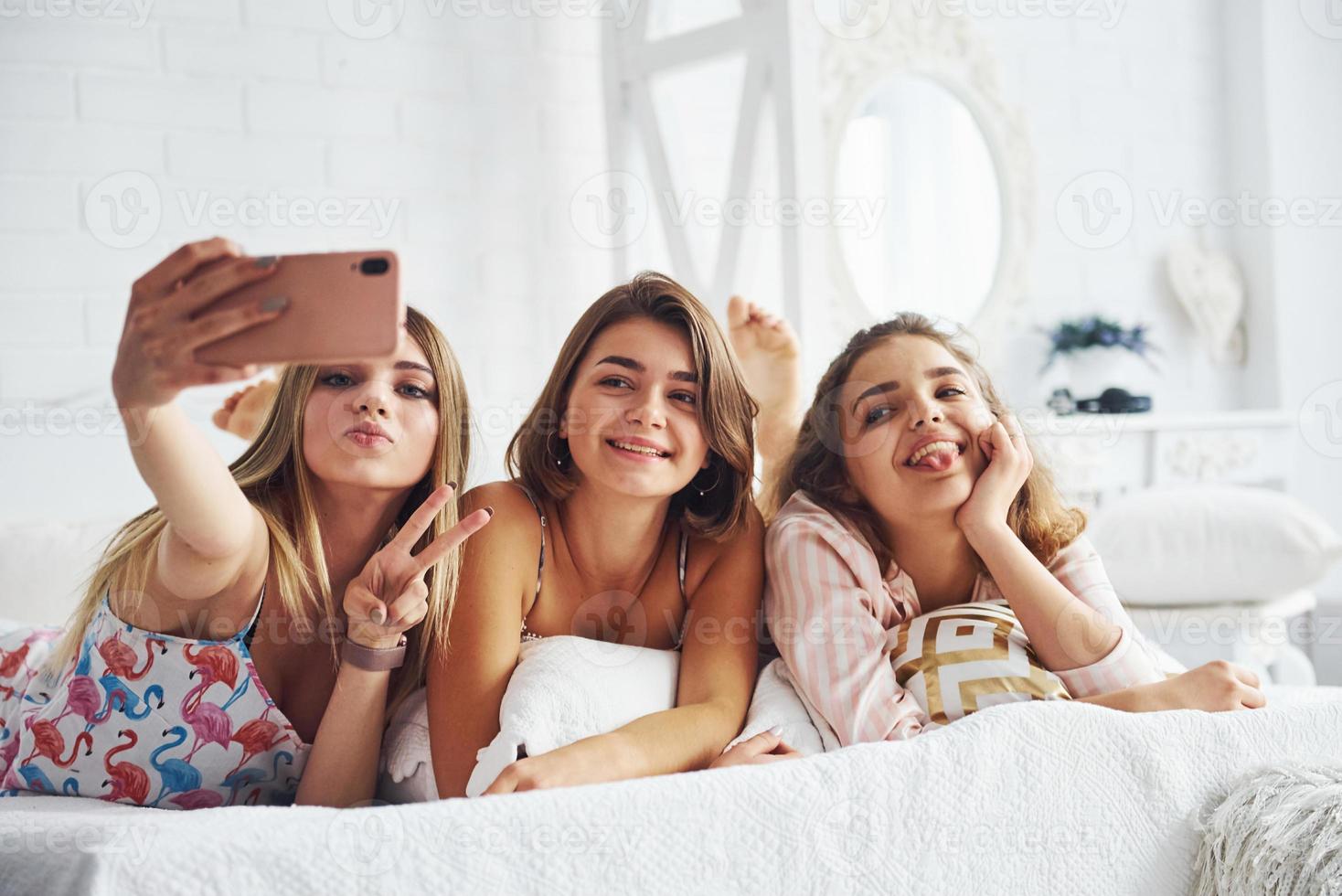 Making selfie by using pink phone. Happy female friends having good time at pajama party in the bedroom photo