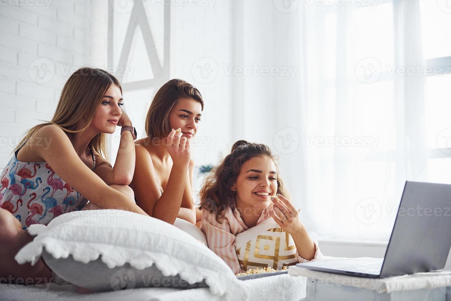 Watching movie on laptop and eating popcorn. Happy female friends having good time at pajama party in the bedroom photo